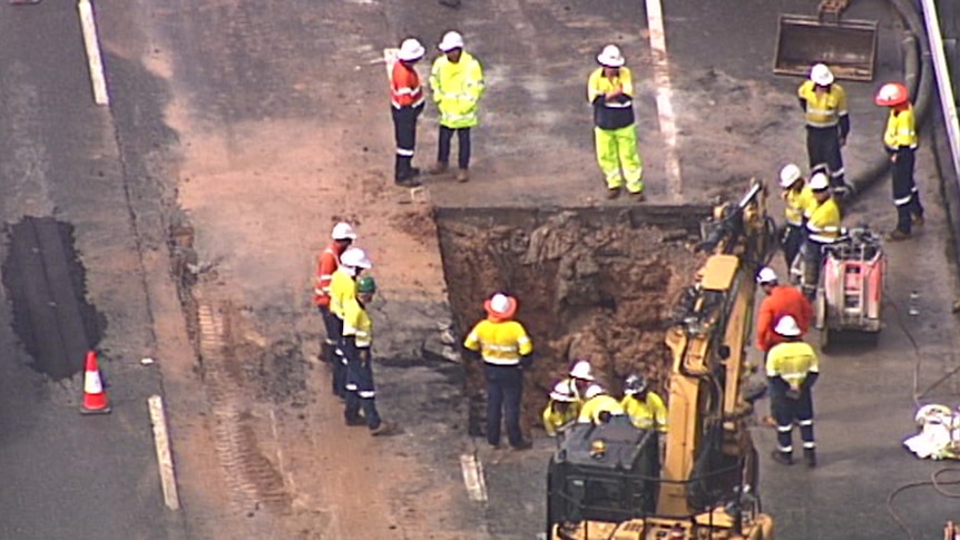 A group of workers in high vis stand around a hold cut in a highway. Earthmoving equipment stands nearby.