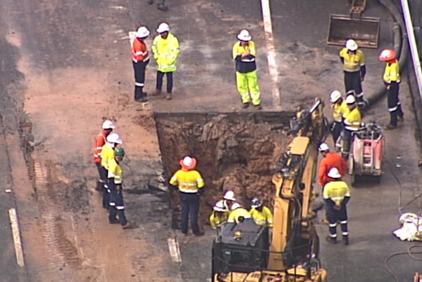 A group of workers in high vis stand around a hold cut in a highway. Earthmoving equipment stands nearby.