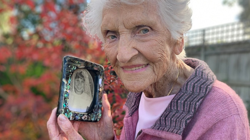 An elderly woman holds a black white photo in a frame while standing in a garden.