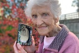 An elderly woman holds a black white photo in a frame while standing in a garden.