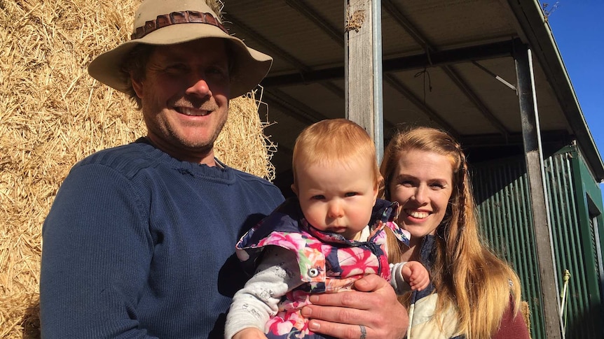 A man in an Akubra-style hat and a woman with long hair smile, holding a young child, in front of hay in a shed.