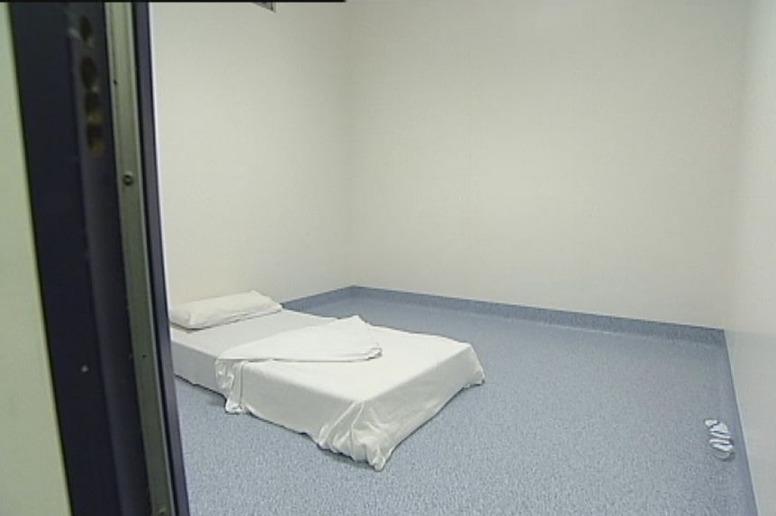 A plain white mattress with a white sheet on a linoleum-looking floor in a room with bare grey walls.