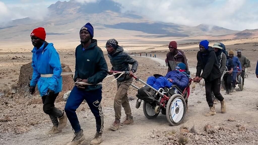 A team of five people push and pull a woman in a wheel chair with long handle bars on a path through an arid desert. 