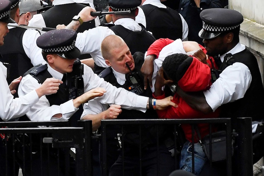 You view a black man in a red hoodie being restrained by a black police officer as his white colleagues rush to restrain him.