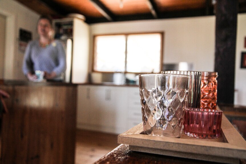 Glasses on a shelf in the foreground with an out-of-focus woman standing behind a kitchen counter in the background.