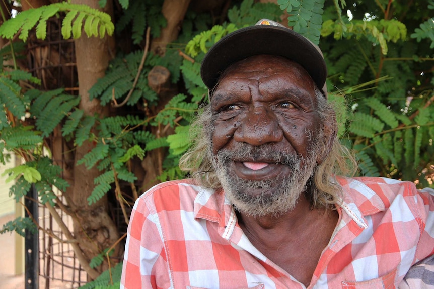 A head and shoulders shot of an older Indigenous man smiling wearing a chequered red shirt and baseball cap outdoors.