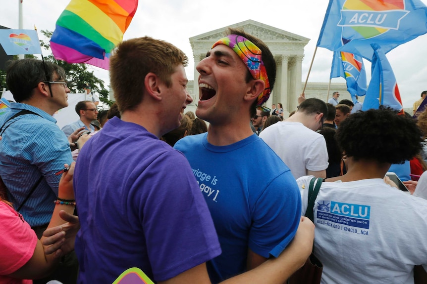 Two men embrace in a huge crowd outside the US Supreme Court. There are several rainbow flags around them