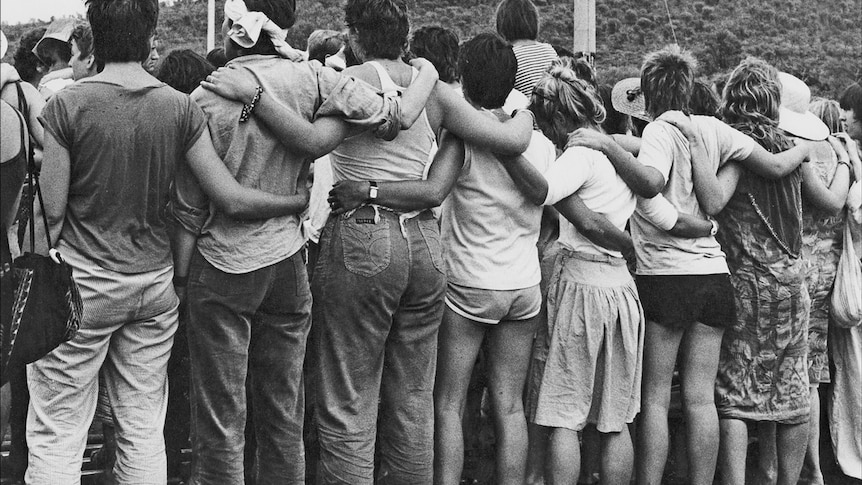Protesters stand linked together at the Pine Gap Women's Peace Camp in 1983, photo taken from behind, bushland in background