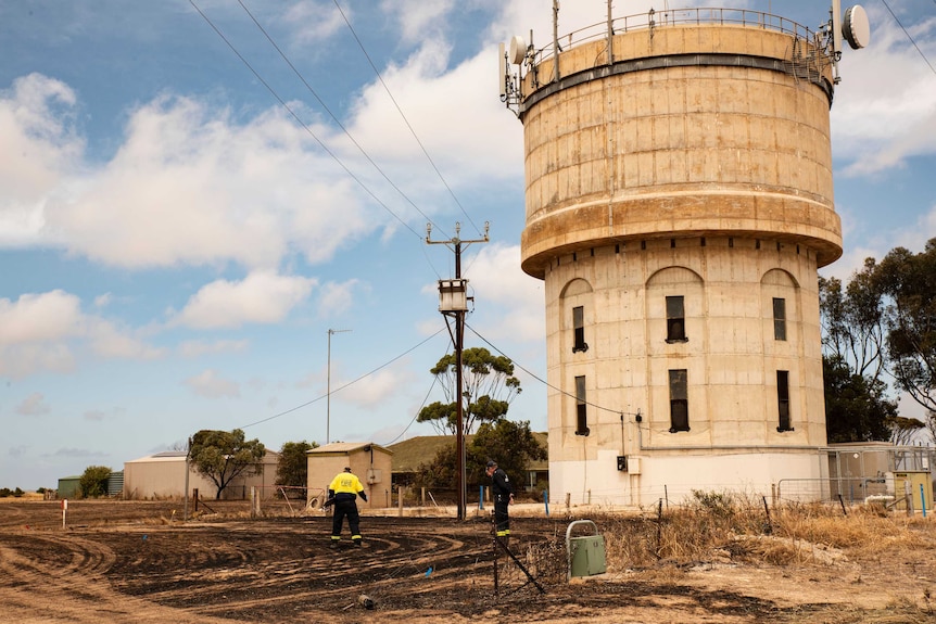 Two men look at burnt ground under a stobie pole with a blackened transformer on top next to a short water tower