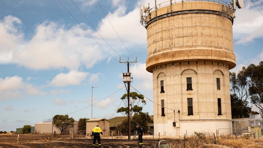Two men look at burnt ground under a stobie pole with a blackened transformer on top next to a short water tower