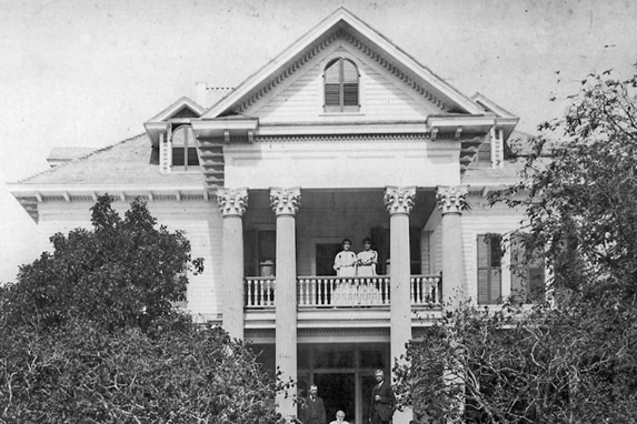 A black and white photo shows two women standing on the upper balcony of a white mansion with greco-style columns.
