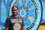 A young Indigenous woman stands in front of a bright blue Indigenous design.