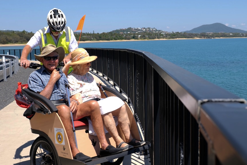 A couple wearing hates, sitting on the bench of the trishaw parked next to a black fence, beside the blue ocean.