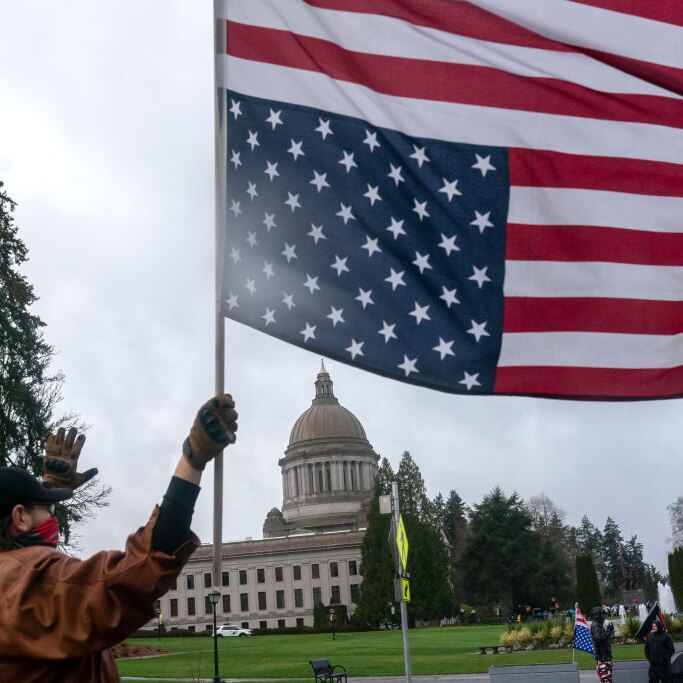 a man wearing a mask and gloves holds the US flag upside down outside the govt offices in WA.