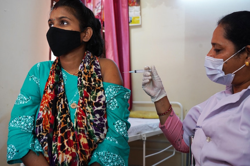 A pregnant woman in a green sari receives a needle into her left arm. 