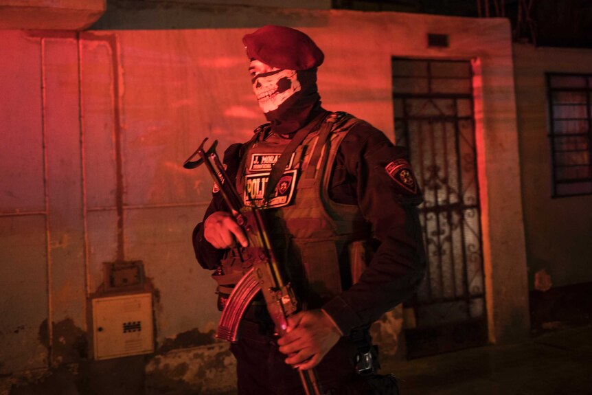 A police officer wearing combat gear, a beret and a bandana with the image of a skull on it stands guard in red street light.
