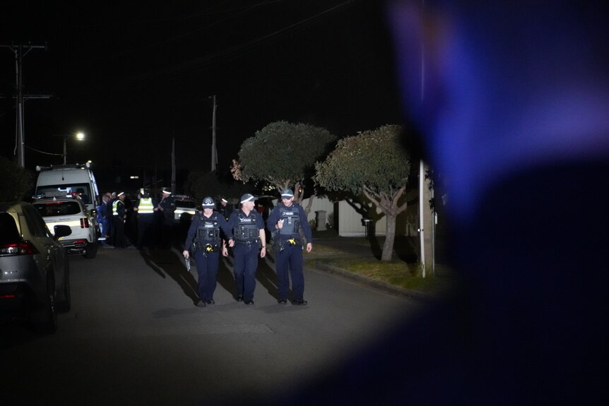 Police officers walk on a dark suburban street. Behind them are emergency vehicles and personnel
