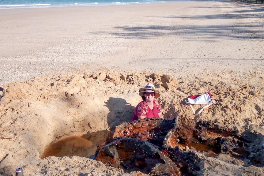 A woman sits in a sand hole with a rusty car body.