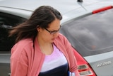 A woman in a pink jacket looks away from the camera as she walks down a suburban street.