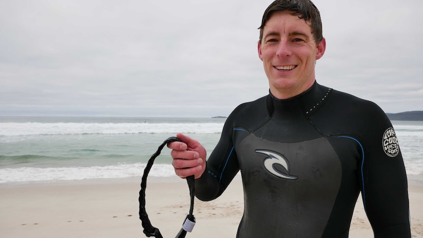 A man in a wetsuit standing on a beach, holding up a tourniquet.