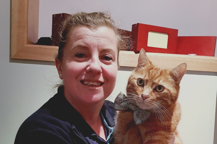 Photograph of a white woman smiling at the camera, holding a ginger cat with a bow tie.