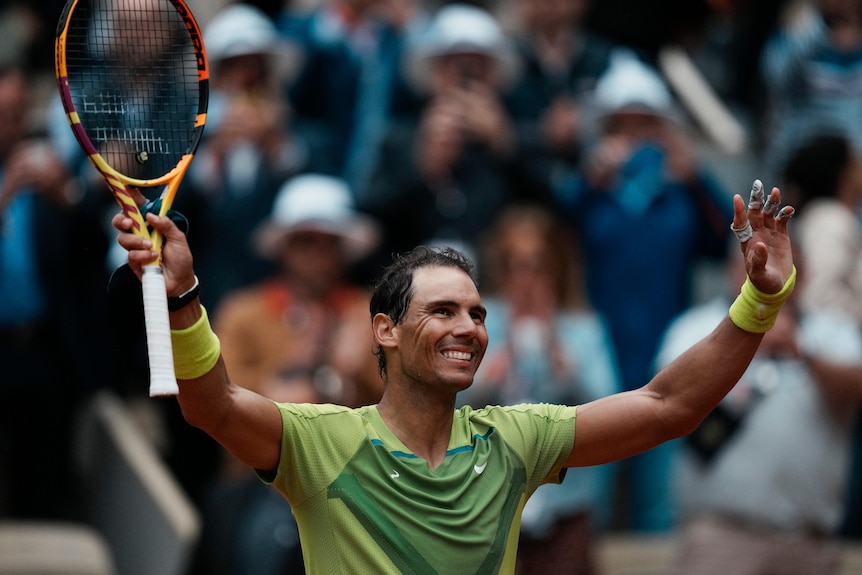 rafael nadal smiles while holding both arms up in the air, one hand holding a tennis racquet, celebrating a win