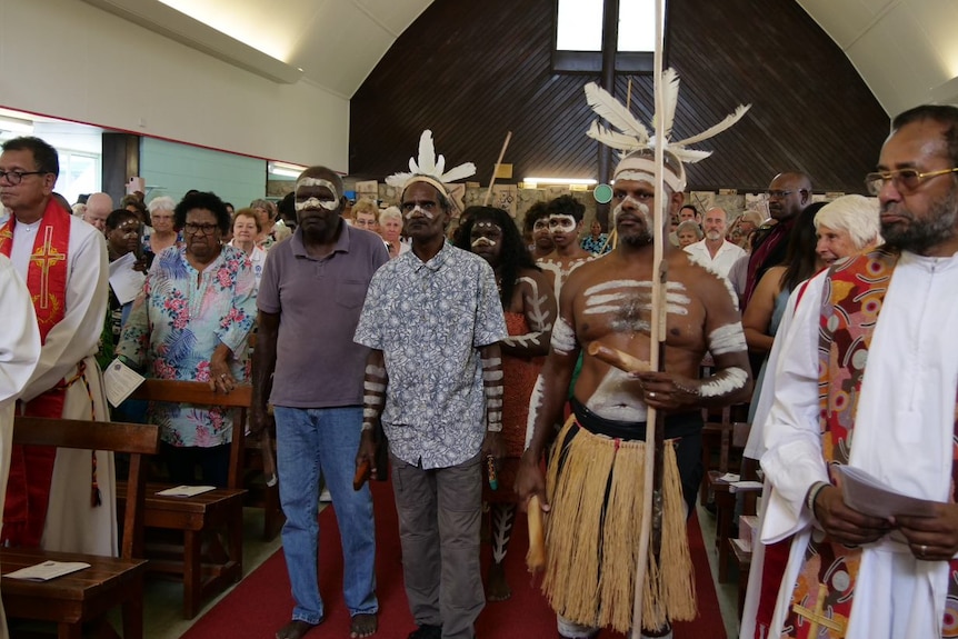 Traditional owners in a church, dressed in ceremonial clothes and markings on body, carrying a staff with feathers.