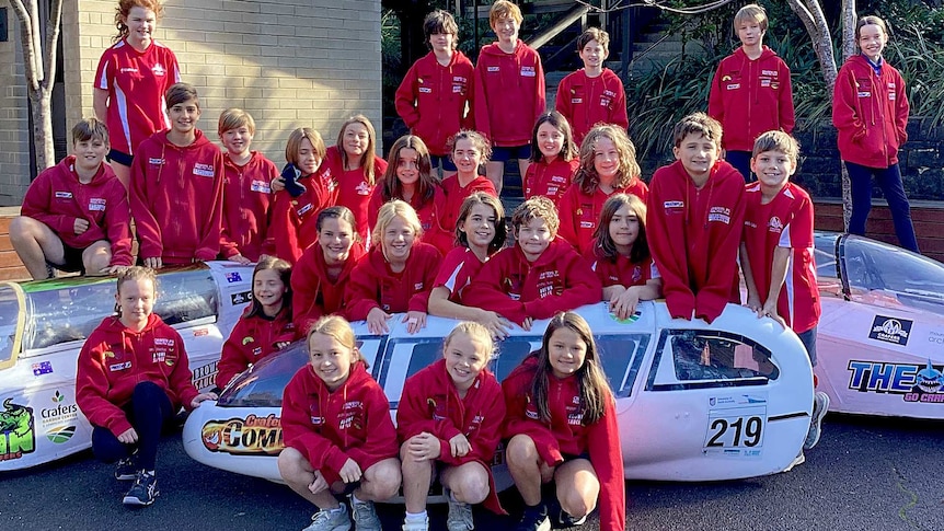 A group of 25 students pose for a photograph amongst three pedal prix vehicles.