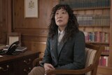 An Asian woman sits in a room with a desk and a bookshelf wearing a grey suit jacket.
