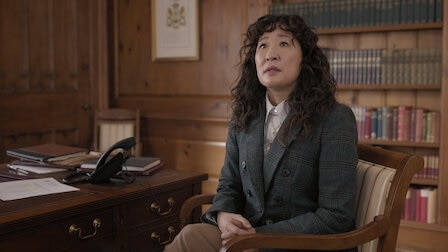 An Asian woman sits in a room with a desk and a bookshelf wearing a grey suit jacket.