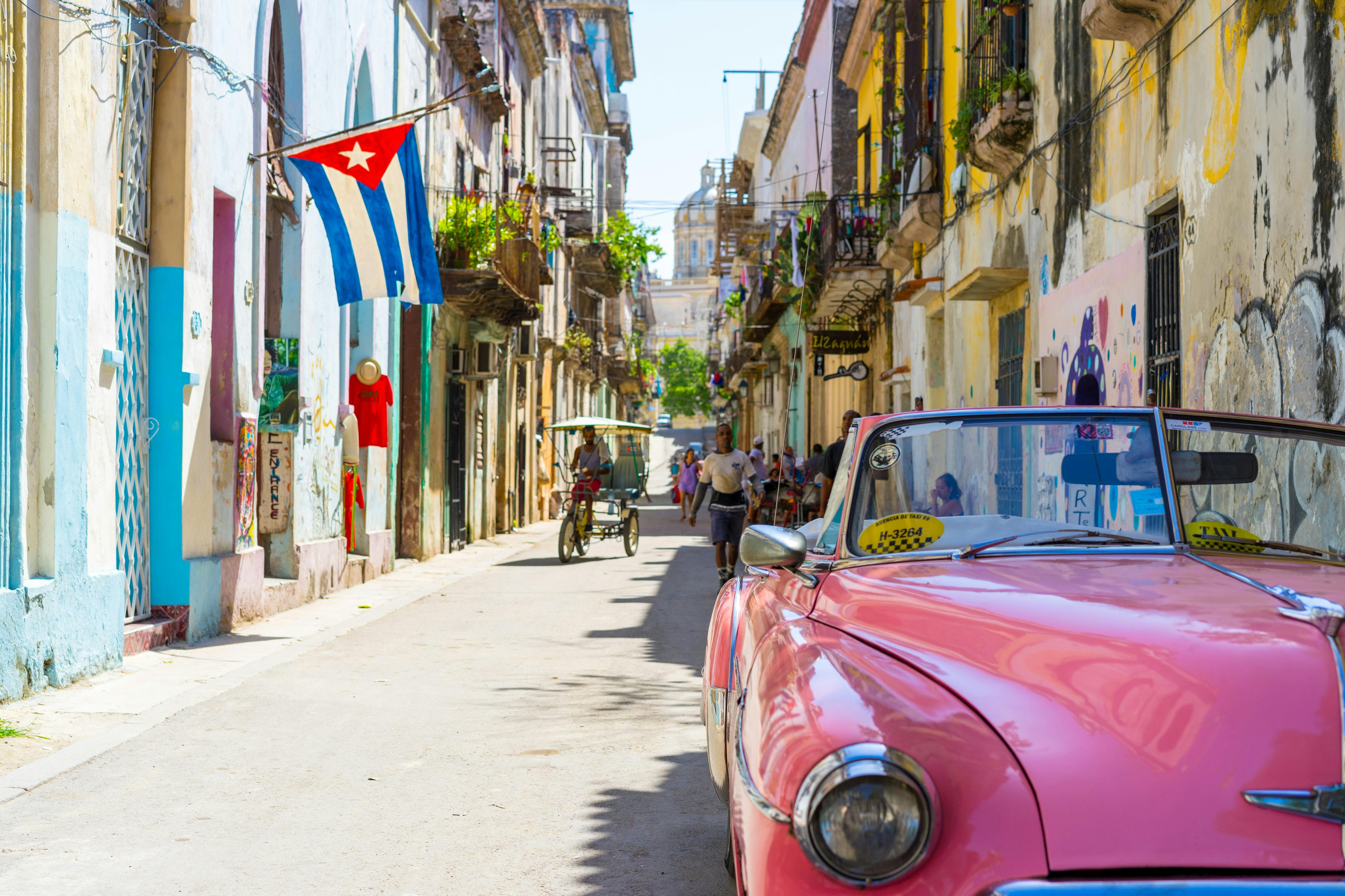  Pink classic car beneath a Cuban flag hung from a pastel balcony on a glowing Havana street alive with colour under midday sun