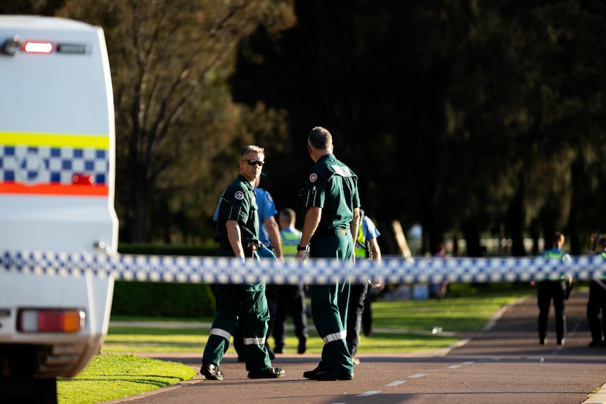 Two St John's WA paramedics look at each other on a footpath with a police car nearby and police tape in the foreground.