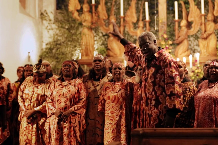 A choir stands singing behind the conductor with his hand raised to the roof