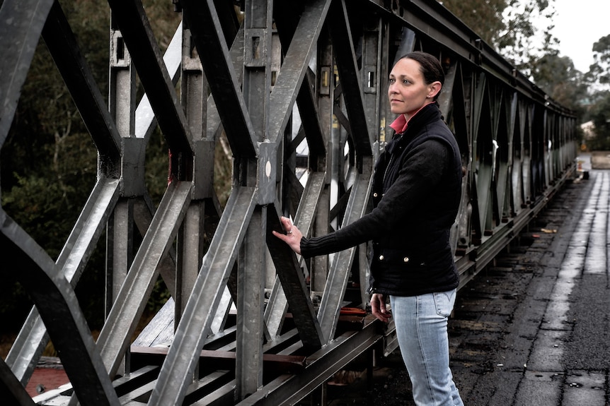 A woman standing on an old damage bridge. She is touching the bridge and looking at damage on the trusses. The bridge is closed.