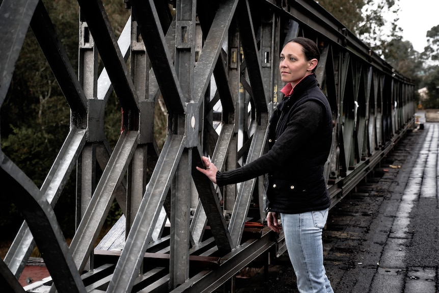 a woman standing on an old damage bridge. She is touching the bridge and looking at damage on the trusses. The bridge is closed 