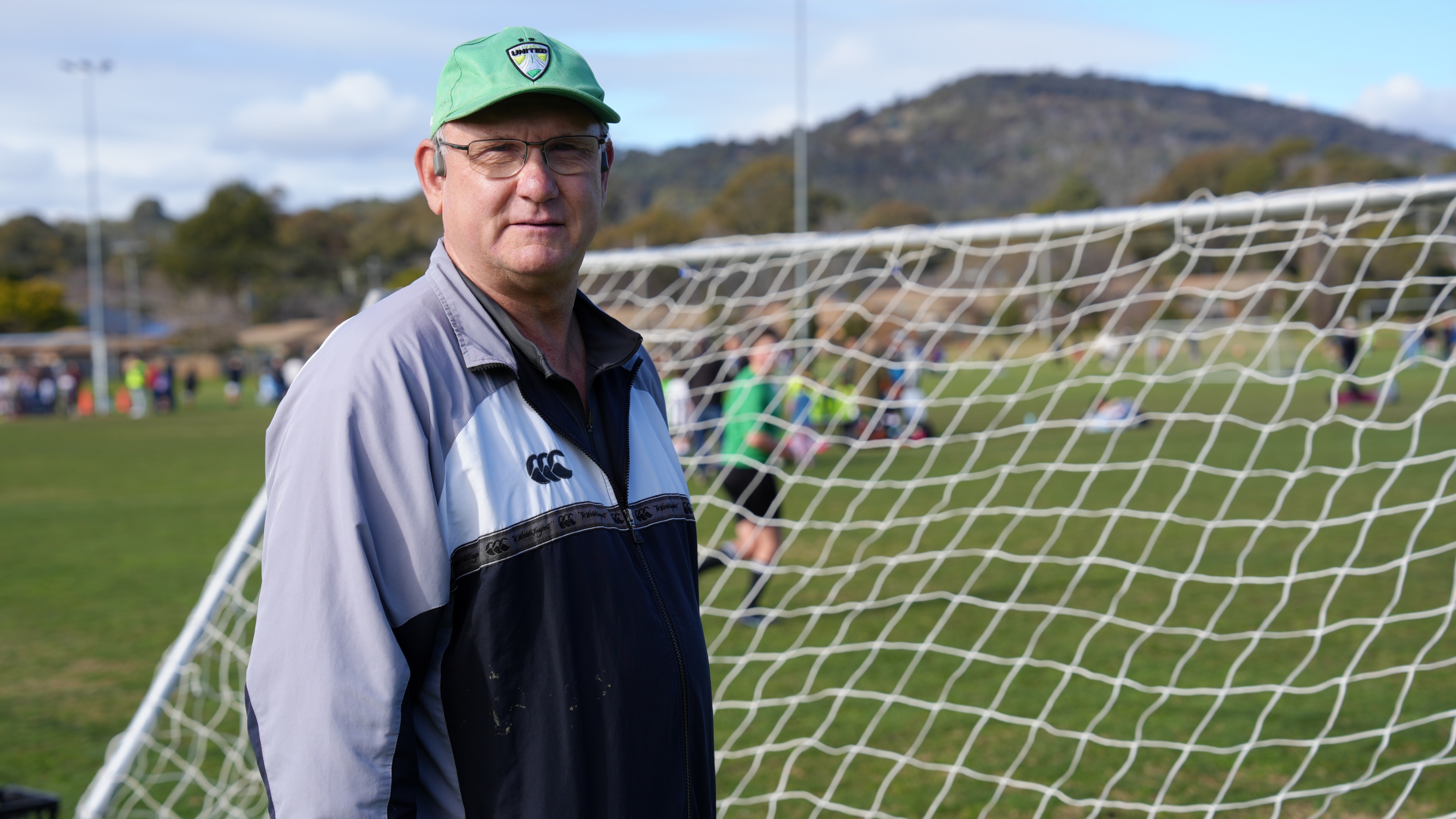A man wearing a green cap stands behind a soccer net on a green playing field.
