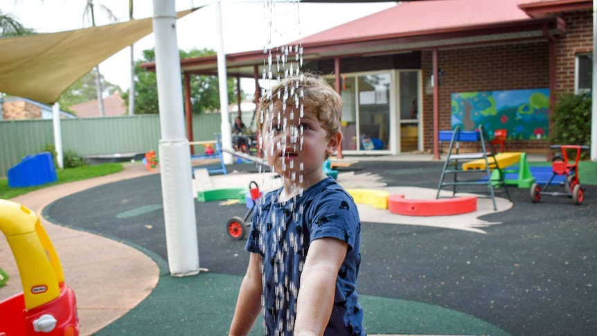 Young boy in blue shirt playing with water at day care centre, colourful toys in background.