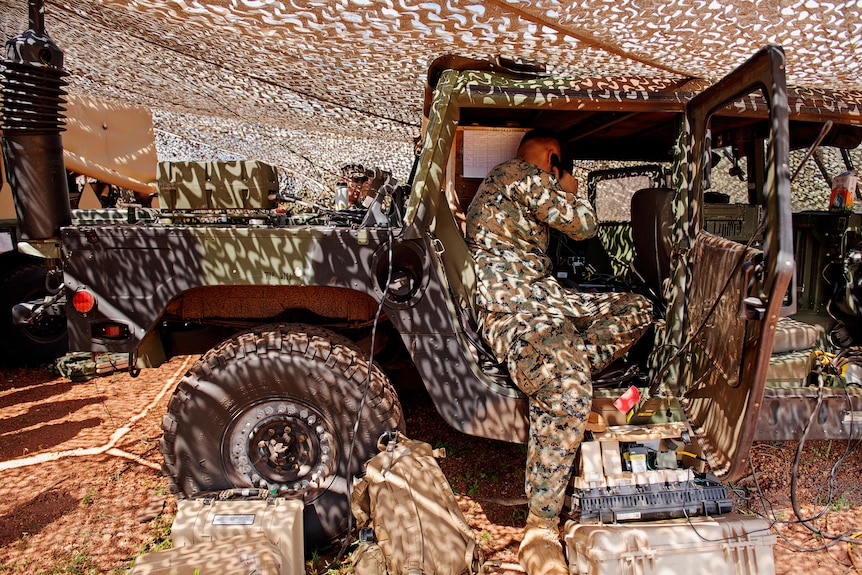 A US Marine in camo fatigues leaning into the open back side door of an army truck parked under a tent, talking on the phone