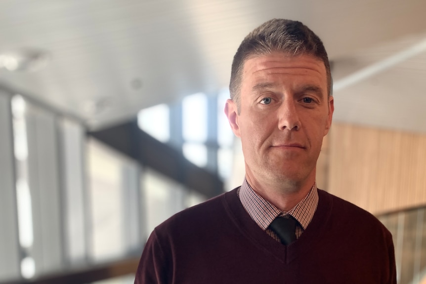 A man in a maroon jumper and tie looks directly into the camera.