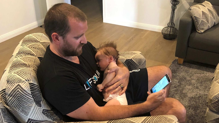 Stuart Horne holds his baby while monitoring his energy use on his smartsphone SmartSun app.
