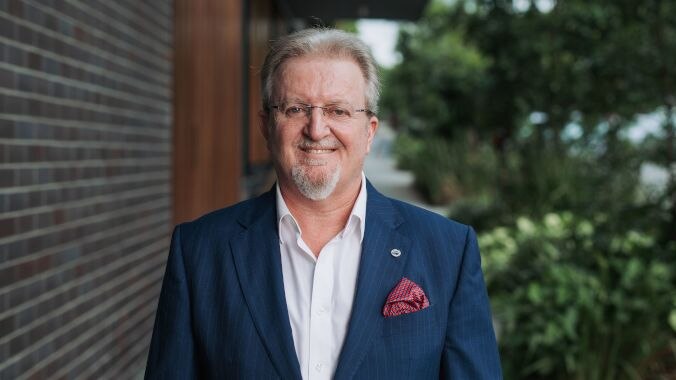 man with grey hair, beard wearing spectacles in blue jacket and red pocket handkerchief smiling in front of building and trees