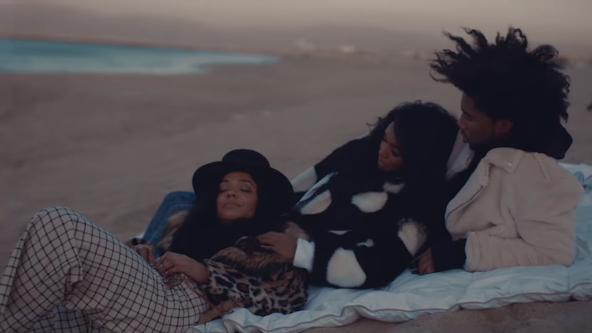 A screenshot from Janelle Monae's Dirty Computer. Janelle lies on a picnic blanket on the beach in between a man and a woman