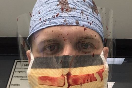 A man wearing a surgical mask and plastic eye guard, both spattered with blood.