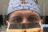 A man wearing a surgical mask and plastic eye guard, both spattered with blood.