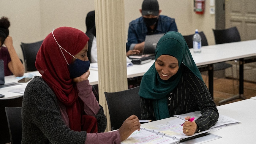 Two women wearing hijab smiling and studying at a table with notebooks open