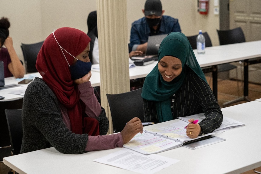 Two women wearing hijab smiling and studying at a table with notebooks open