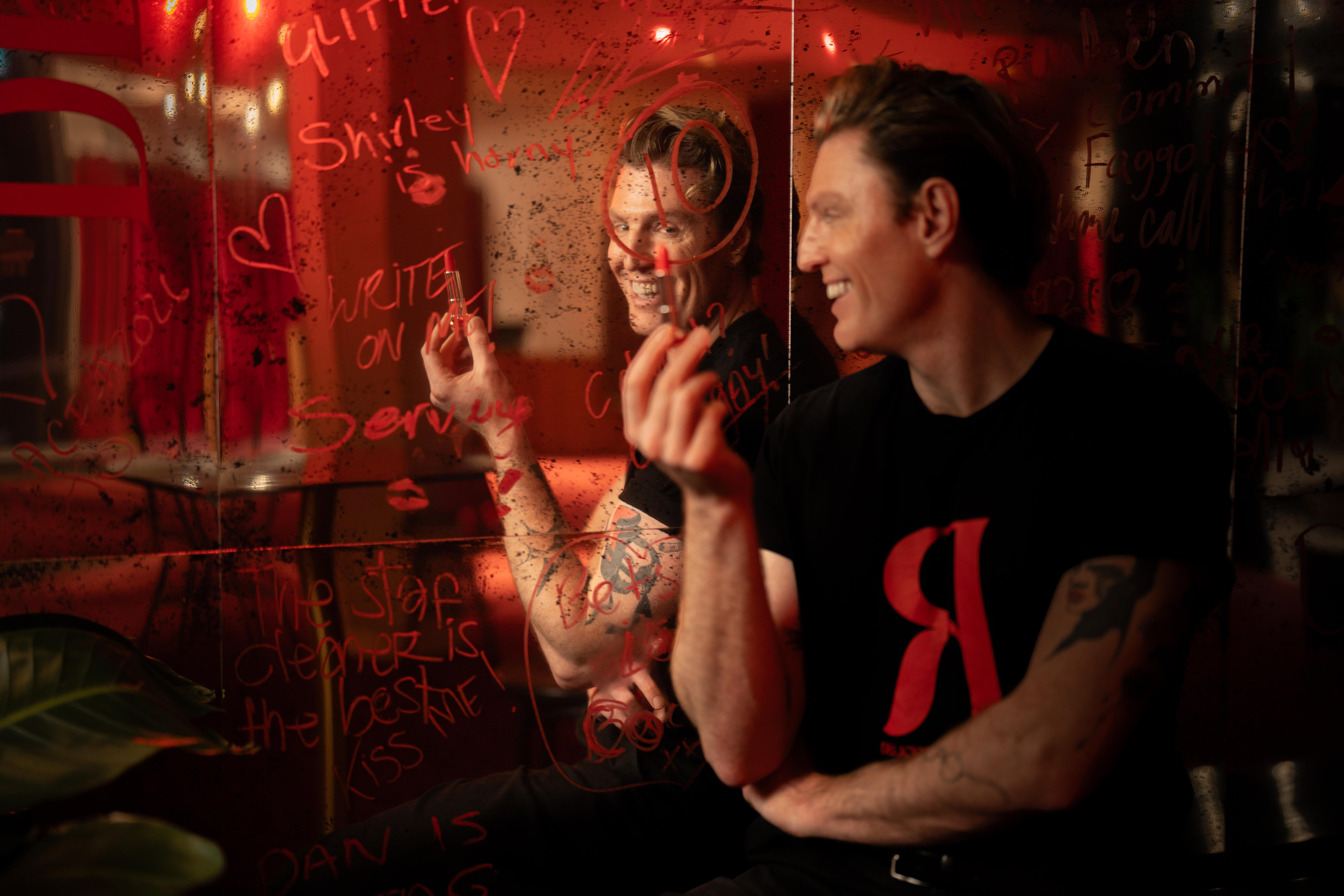 Reuben Kaye, 41, leans back against a mirror, covered in lipstick scrawls, smiling widely, holding a lipstick tube in his hand.