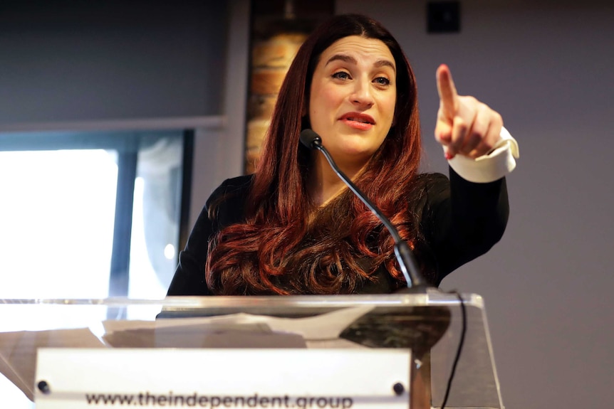 A woman stands at a lectern while pointing her finger at someone in the crowd behind the camera.