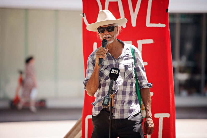 A man in a brimmed hat, sunglasses and flannelette shirt speaking into a microphone, in front of a sign, at a protest.