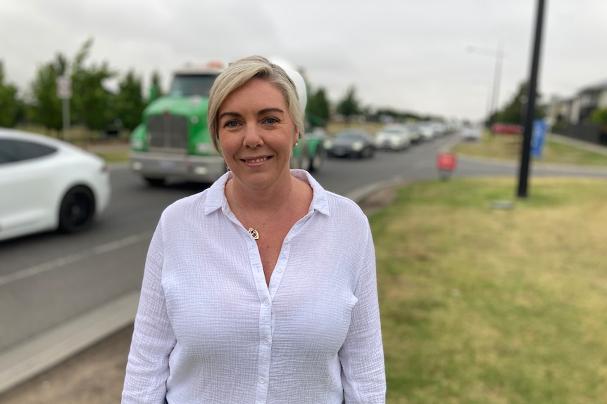 Ally Watson, wearing a white shirt and standing near a busy road which has trucks and cars driving on it.